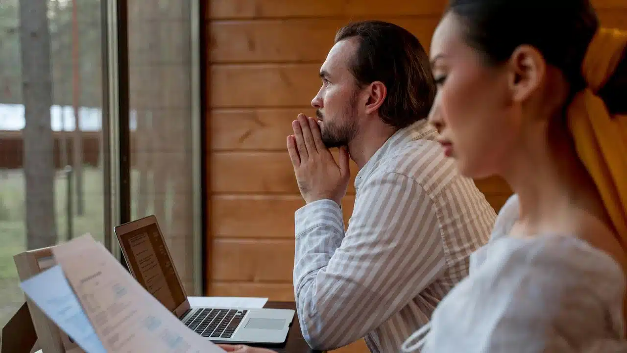 A man and woman sitting by a window, appearing thoughtful while reviewing documents.