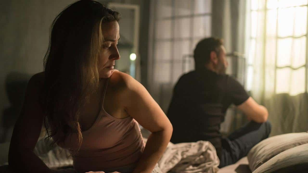 An upset mid-adult woman with her husband in bed behind her.