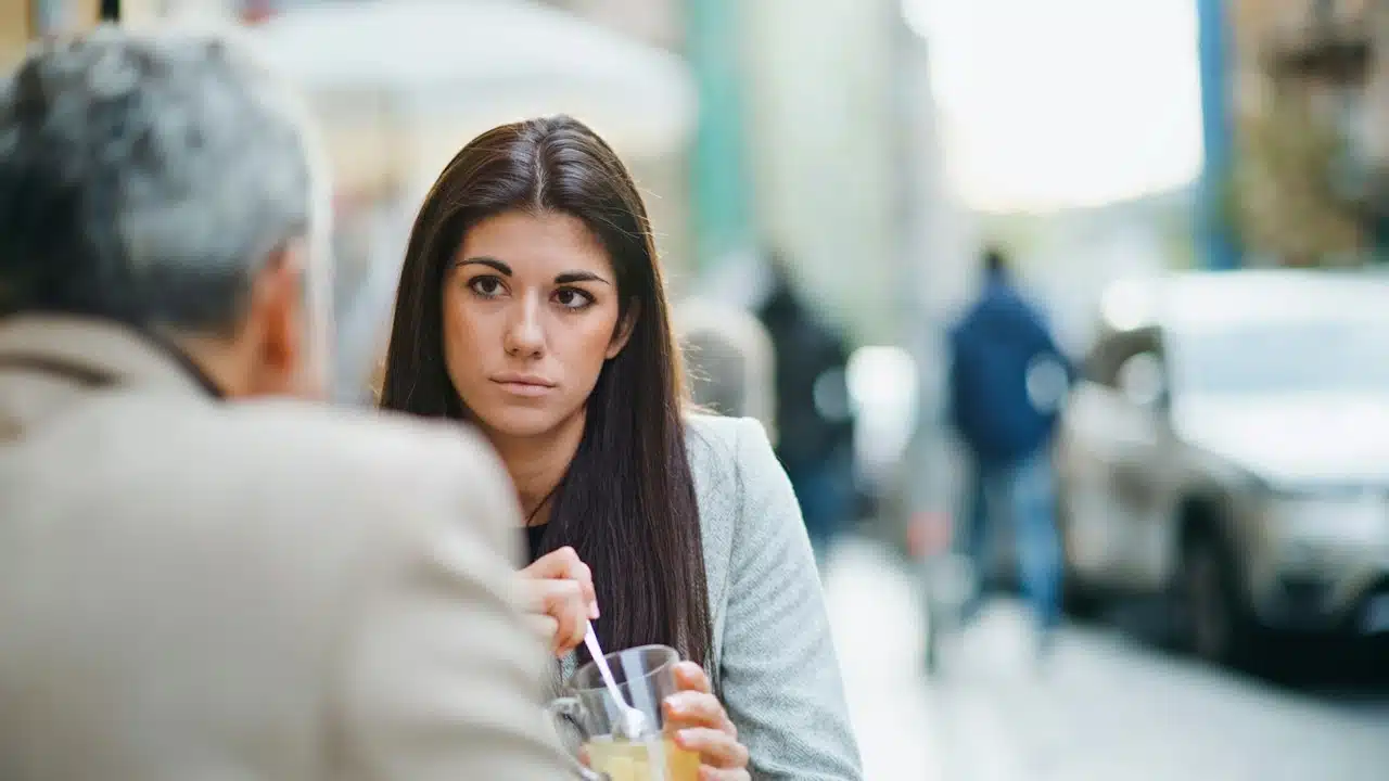 A woman with long dark hair sits at an outdoor cafe, looking seriously at a person across from her while stirring a glass of tea.