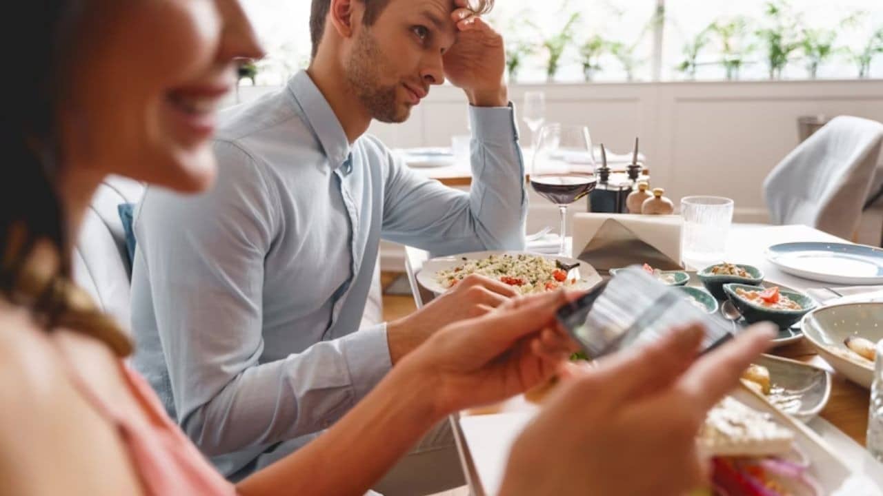 A smiling woman serving dinner  and taking pictures of the food while her partner looks uninterested