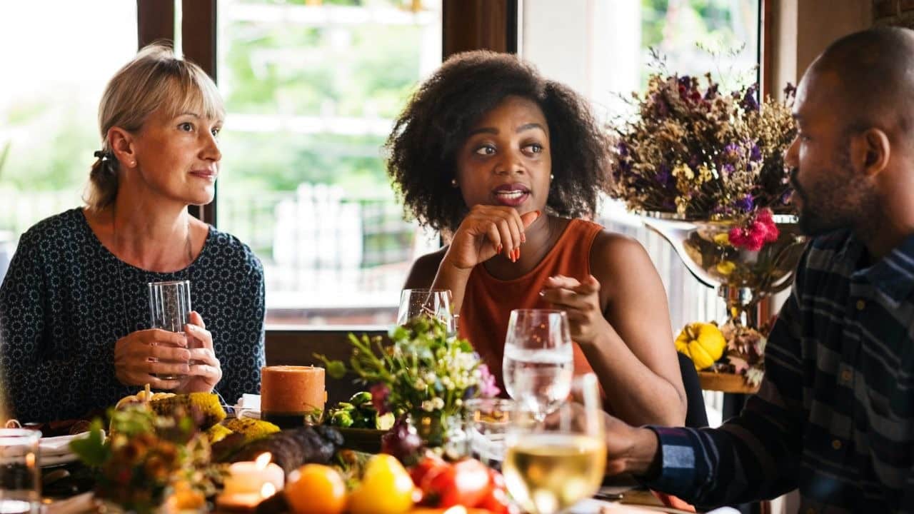 Three people sit around a dinner table with food and drinks, engaged in conversation.