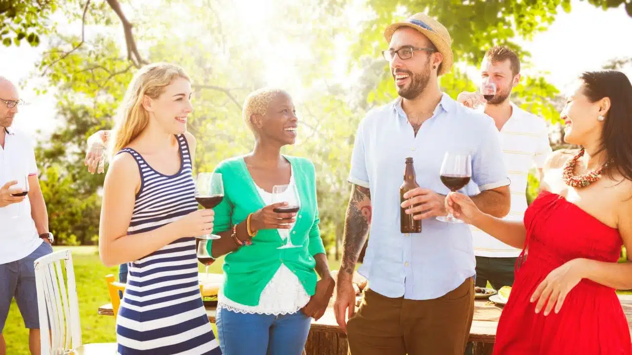 A diverse group of friends laughs and talks together outdoors while holding various drinks.