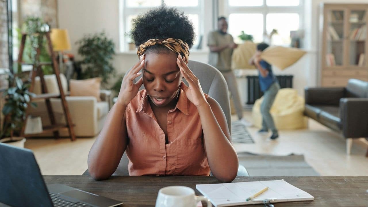 A woman holds her head at a desk while two people play in the background.