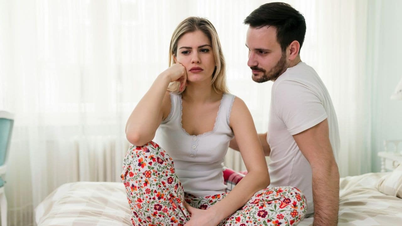 A woman in floral pants sits on a bed while a man looks at her.