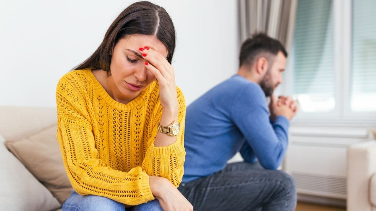 A woman in a yellow sweater touches her forehead while a man sits behind her.