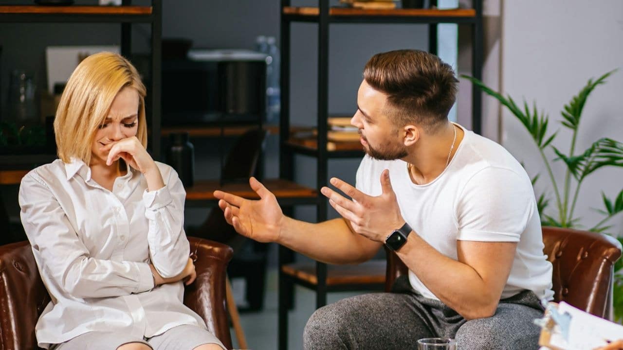 A man in a white t-shirt gestures while talking to a woman who is crying.