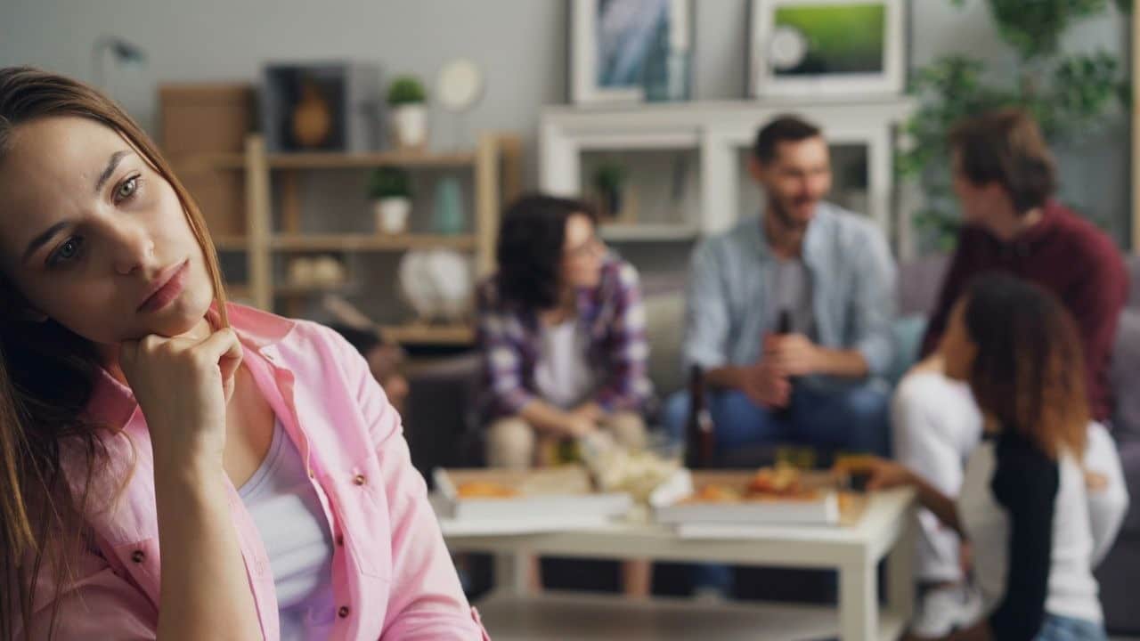 A woman rests her chin on her hand while people talk in a blurred background.