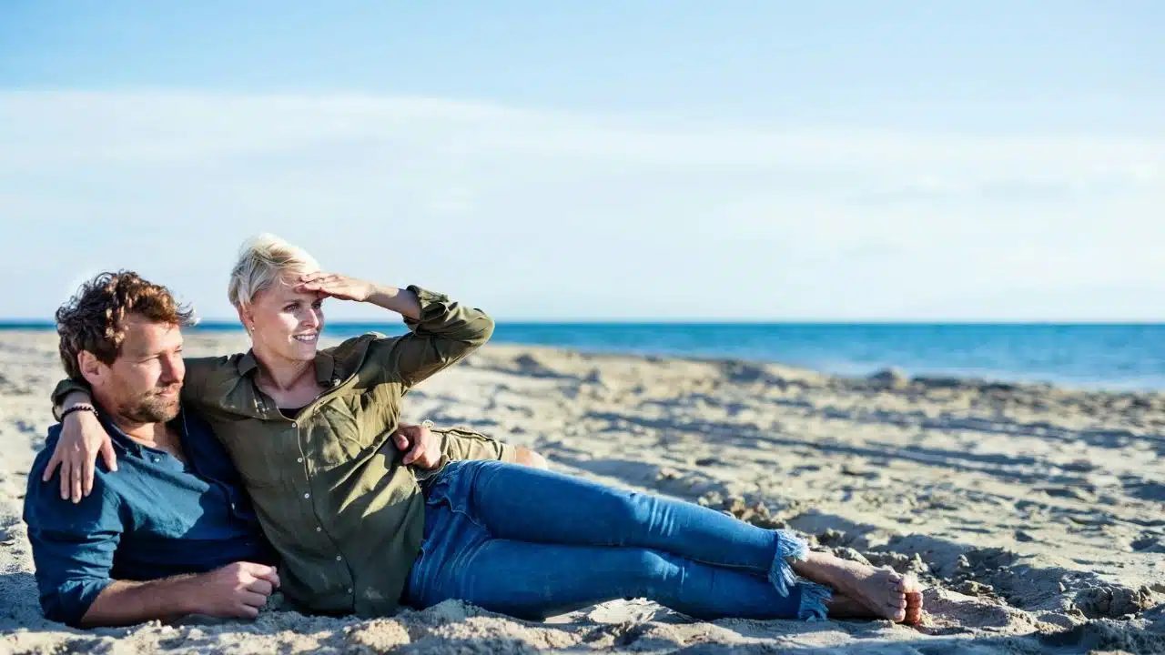 A couple reclines on a sandy beach, looking toward the ocean horizon on a sunny day.