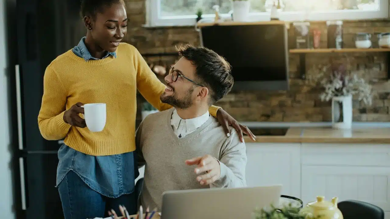 A woman stands with a mug, smiling down at a seated man working on a laptop.