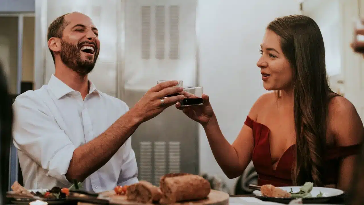 A laughing man and a smiling woman toast with small glasses during a meal together.