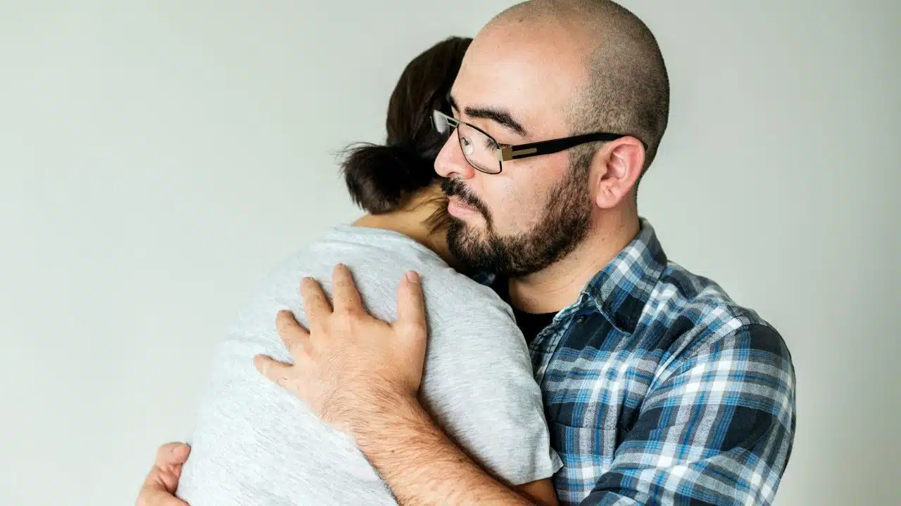 A man wearing glasses and a plaid shirt embraces another person in a close, firm hug.