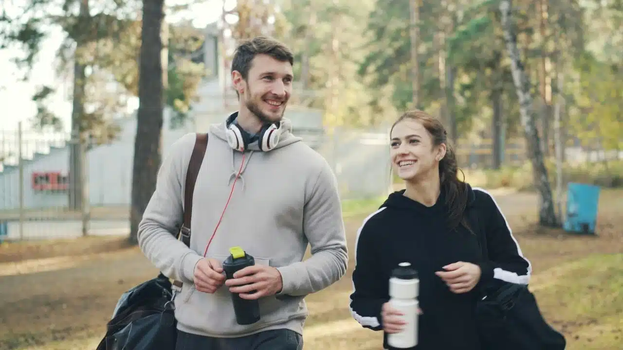 A smiling man and woman walk through a park carrying gym bags and water bottles.