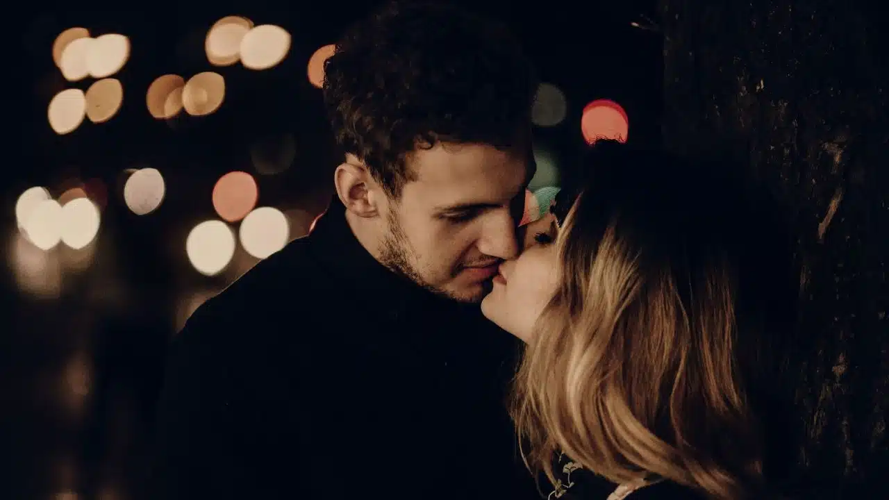 A man and woman share an intimate kiss at night against a backdrop of blurry lights.