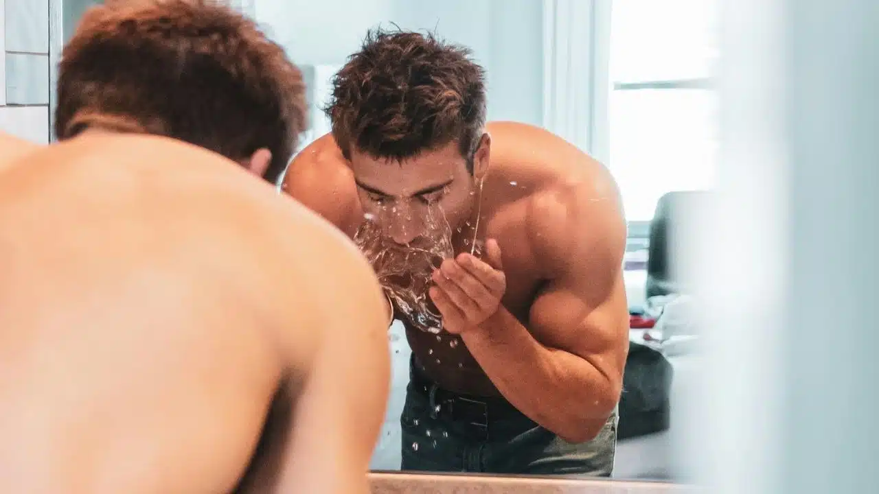 A shirtless man splashes water on his face while looking into a bathroom mirror.
