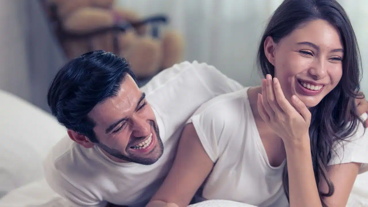 A man and woman in white shirts lie down together while laughing and smiling brightly.