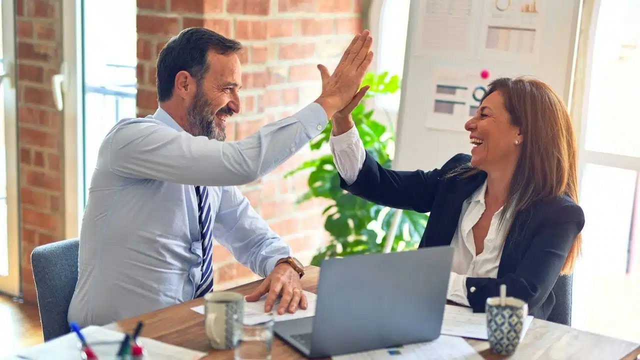 A man and woman in professional attire laugh while giving each other a high five.