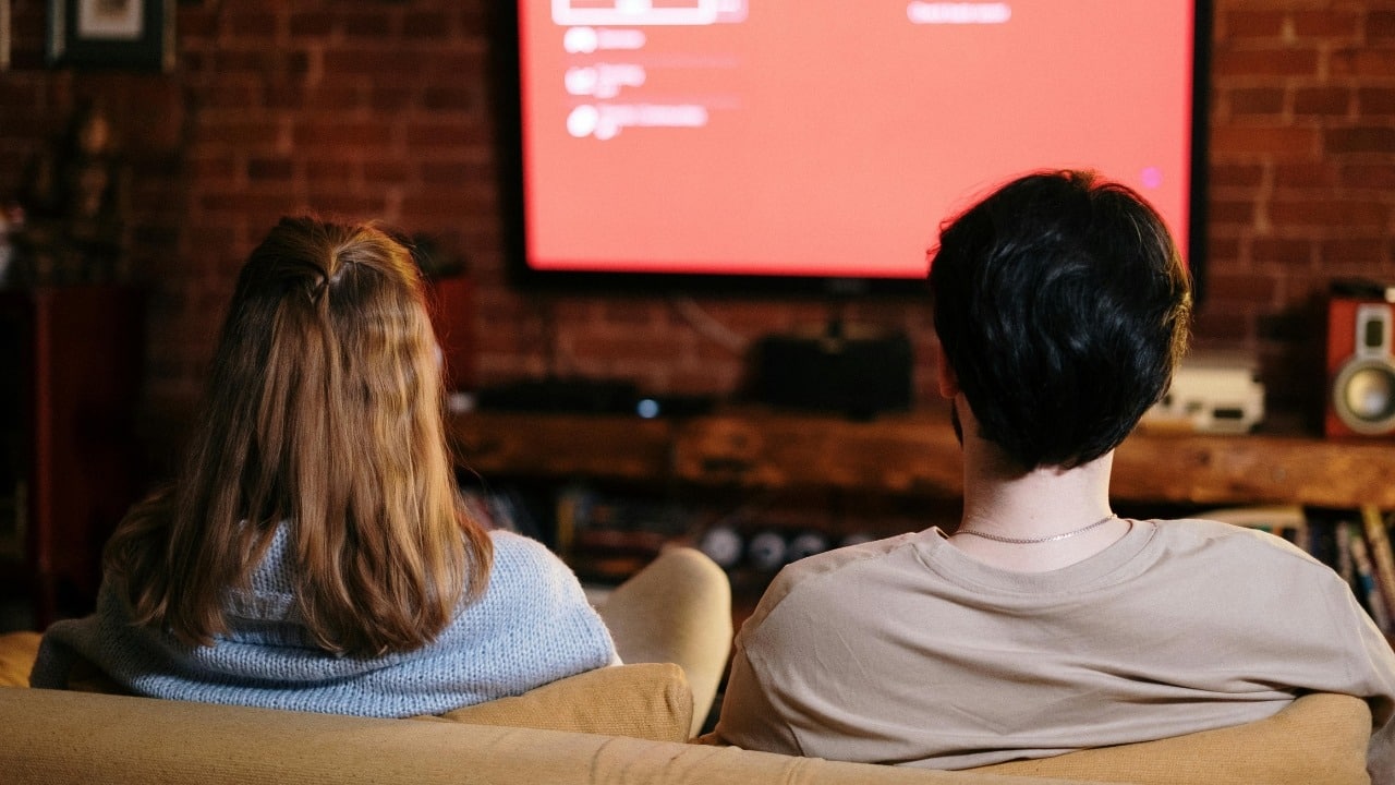 A man and woman sit on a couch viewed from behind watching a large television.