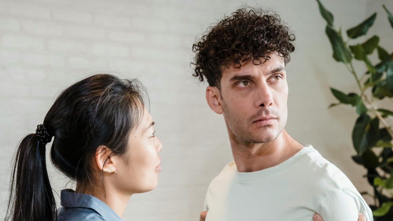 A man looks away with a serious expression while a woman stands facing him.