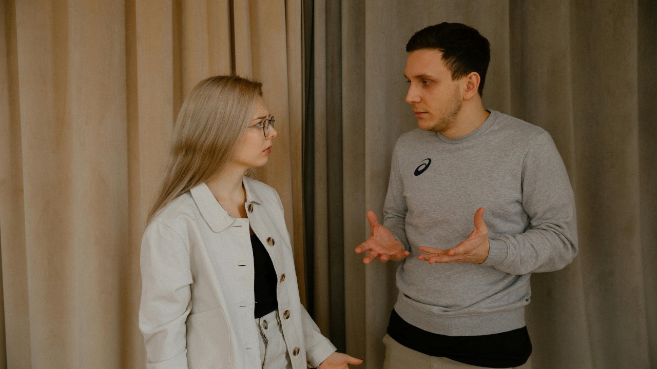 A man gestures with open hands while speaking to a woman in a beige jacket.