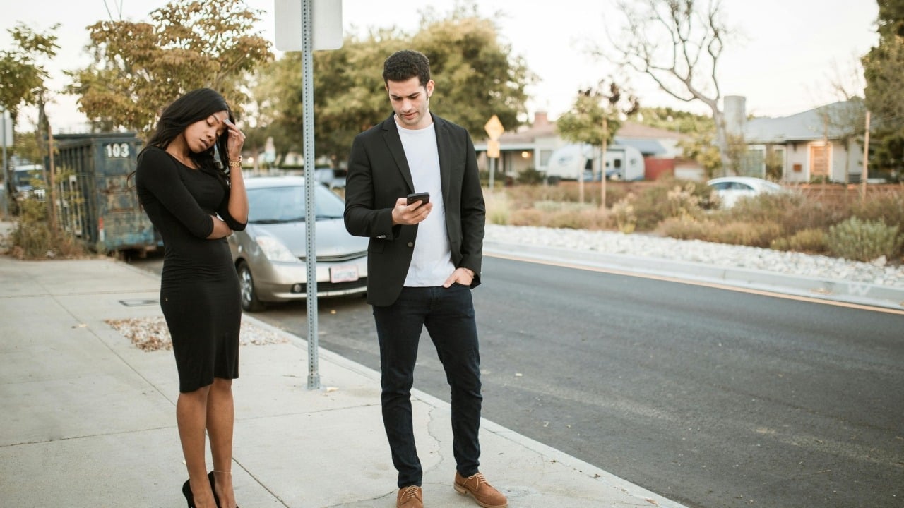 A man looks at his phone while a woman stands nearby with her hand up.