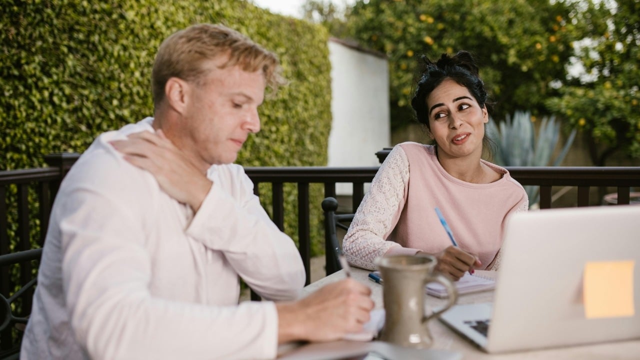 A man and woman sit at an outdoor table with a laptop and writing notebooks.