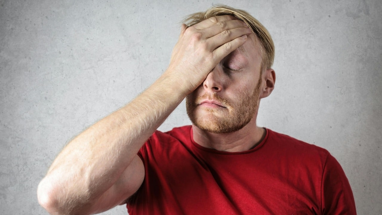 A man in a red t-shirt covers his eyes with his hand against a gray wall.