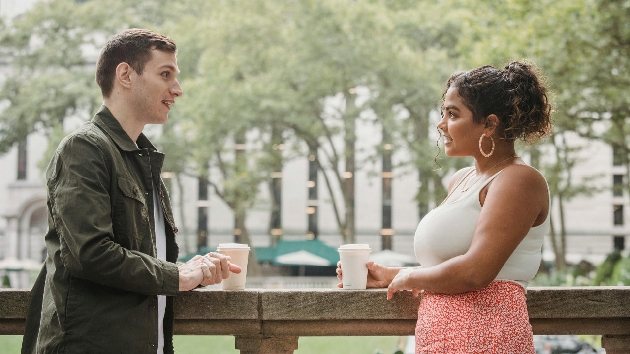 A man and woman stand by a stone railing outdoors holding paper coffee cups.