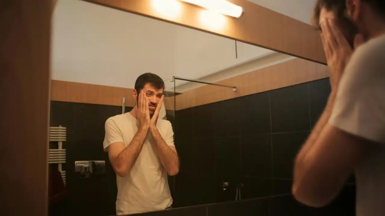 A man in a white t-shirt looks into a bathroom mirror while touching his face.