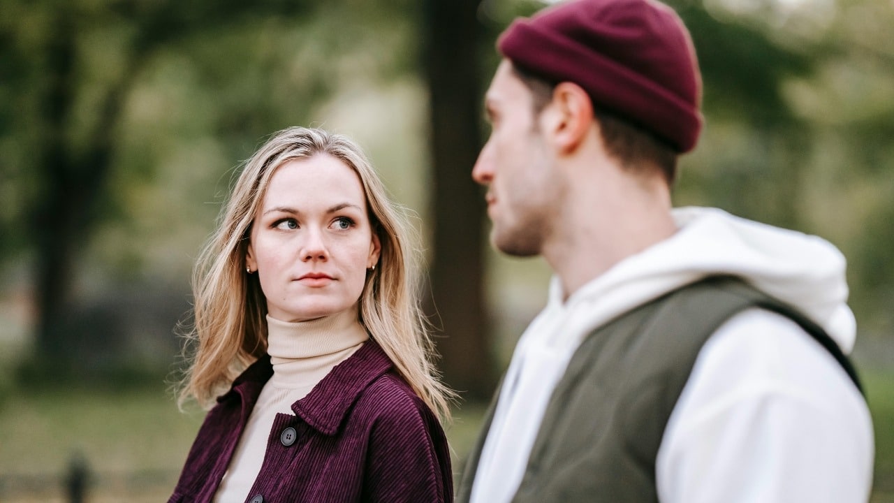 A blonde woman looks toward a man wearing a maroon beanie and a green vest outdoors.