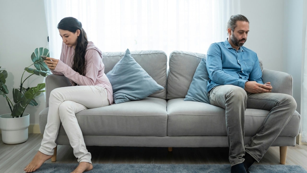 A man and woman sit back-to-back on a gray couch while looking at their phones.