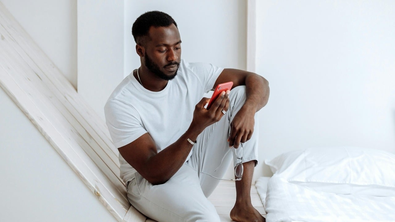 A man in a white t-shirt sits on a bed looking at a red smartphone.