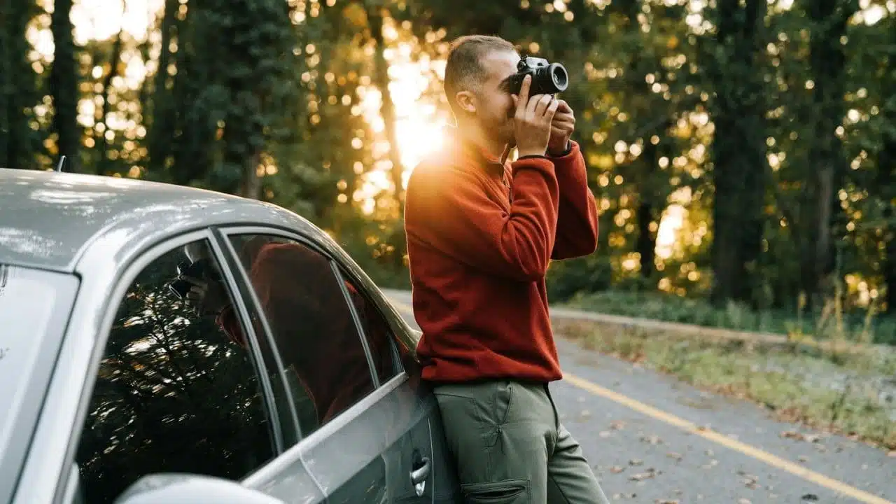 A man in a red sweater leans on a car and takes a photo.