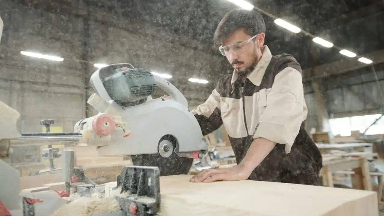 A man in safety glasses uses a miter saw to cut a wooden plank.