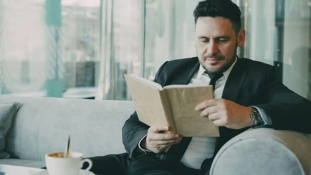 A man in a suit and tie sits on a couch reading a hardcover book.
