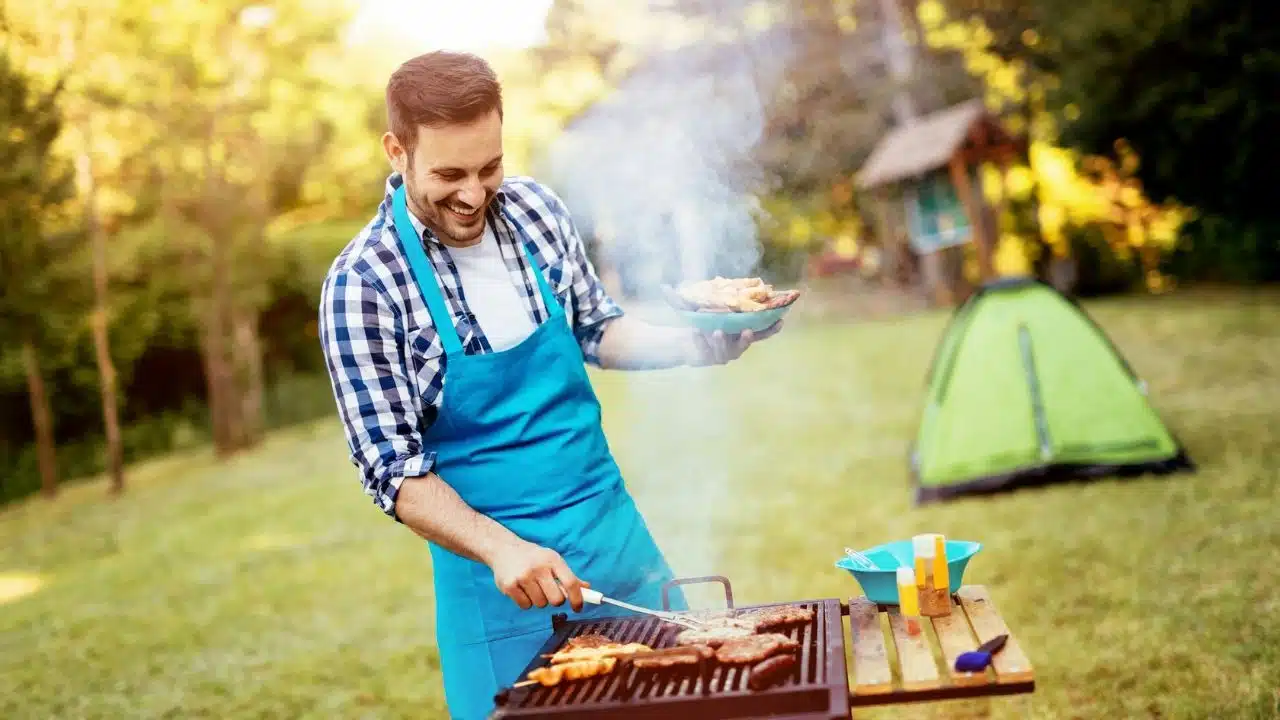 A man in a blue apron smiles while grilling food outdoors near a green tent.