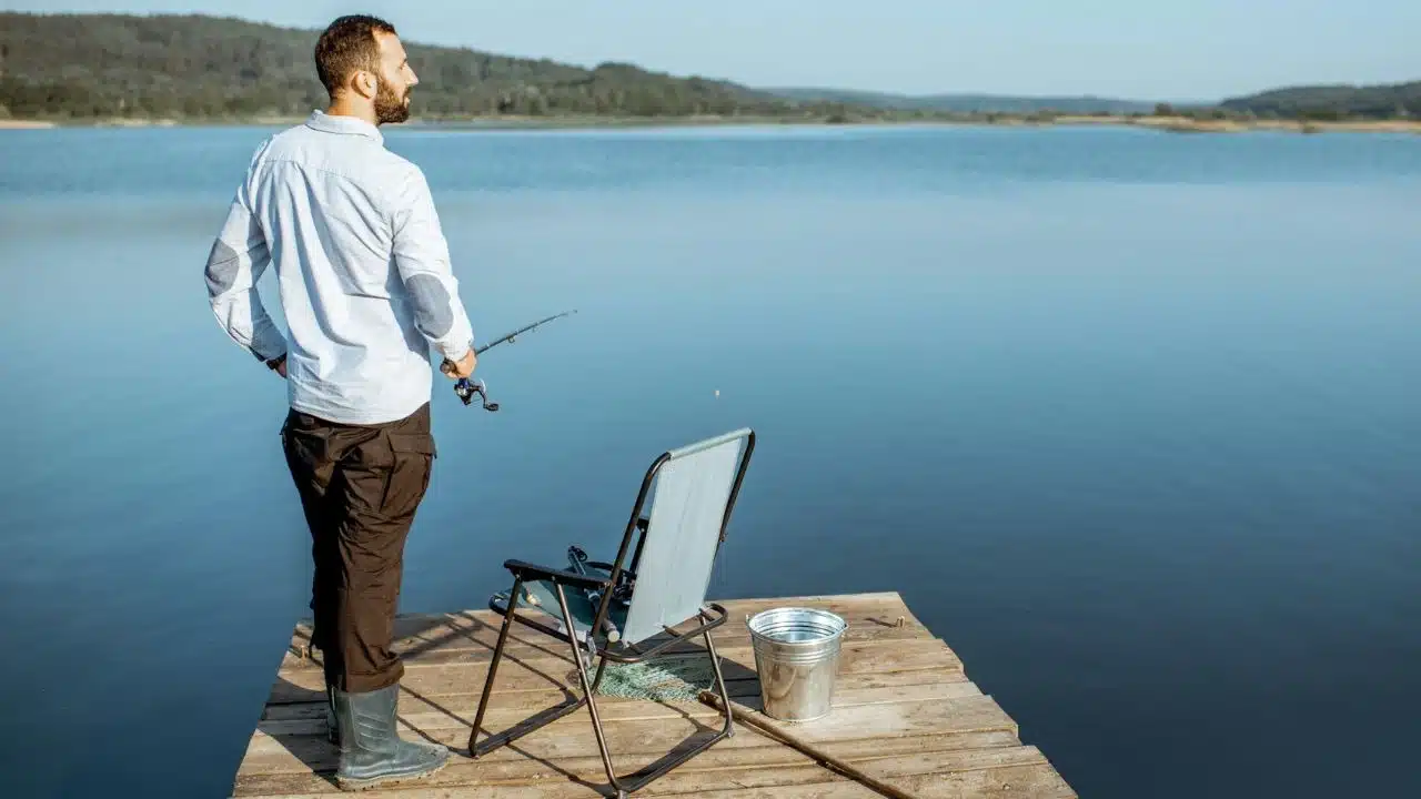 A man stands on a wooden dock holding a fishing rod overlooking a calm lake.