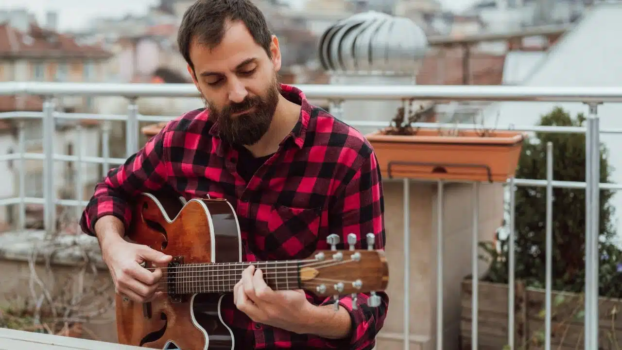 A bearded man in a plaid shirt plays an acoustic guitar on a balcony.