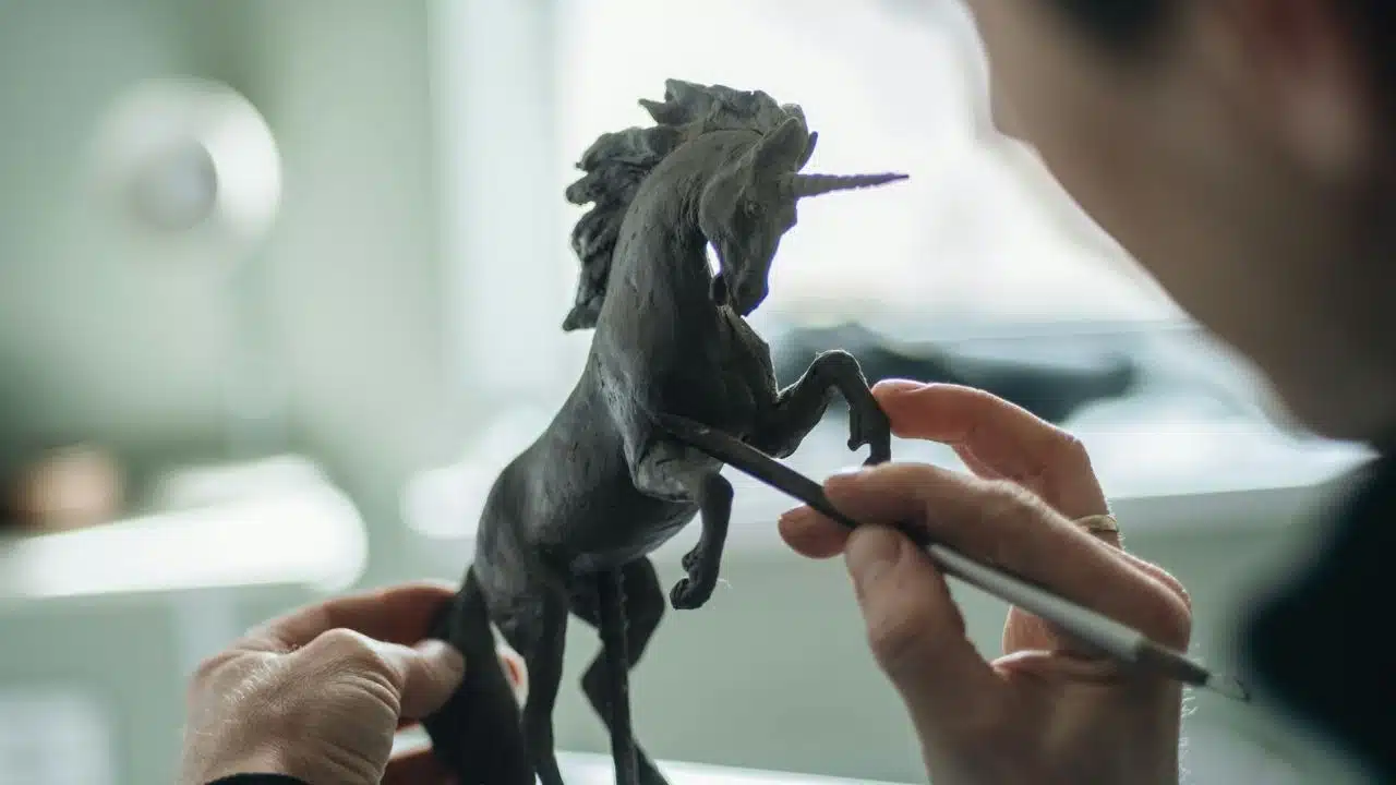 Hands use a small tool to detail a dark, rearing unicorn sculpture on a table.