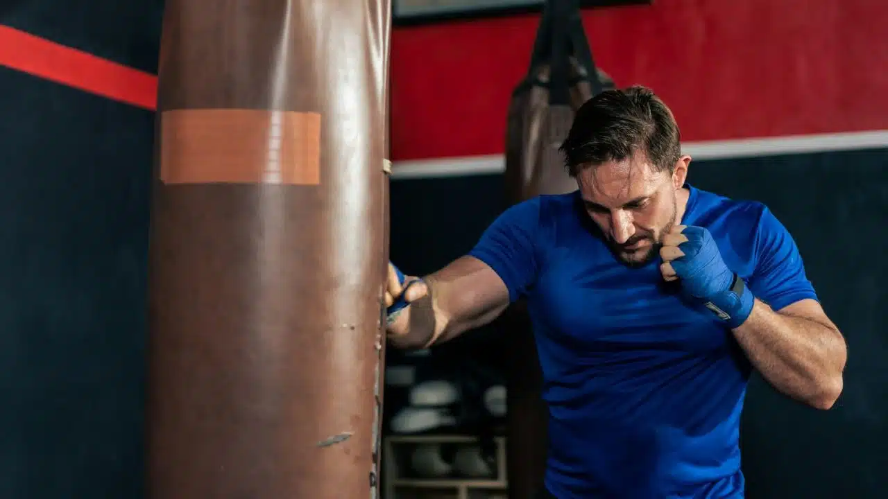 A man with blue hand wraps punches a large brown heavy bag in a gym.