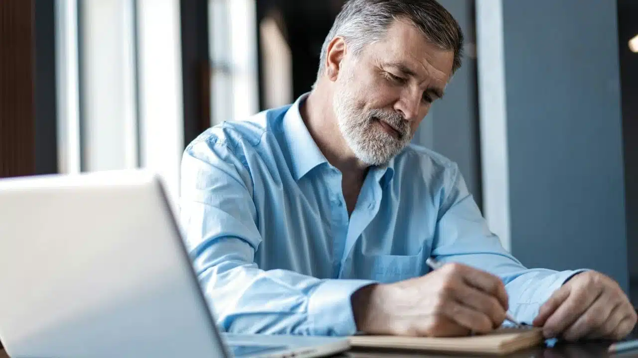 A bearded man in a blue shirt writes in a notebook beside a laptop.