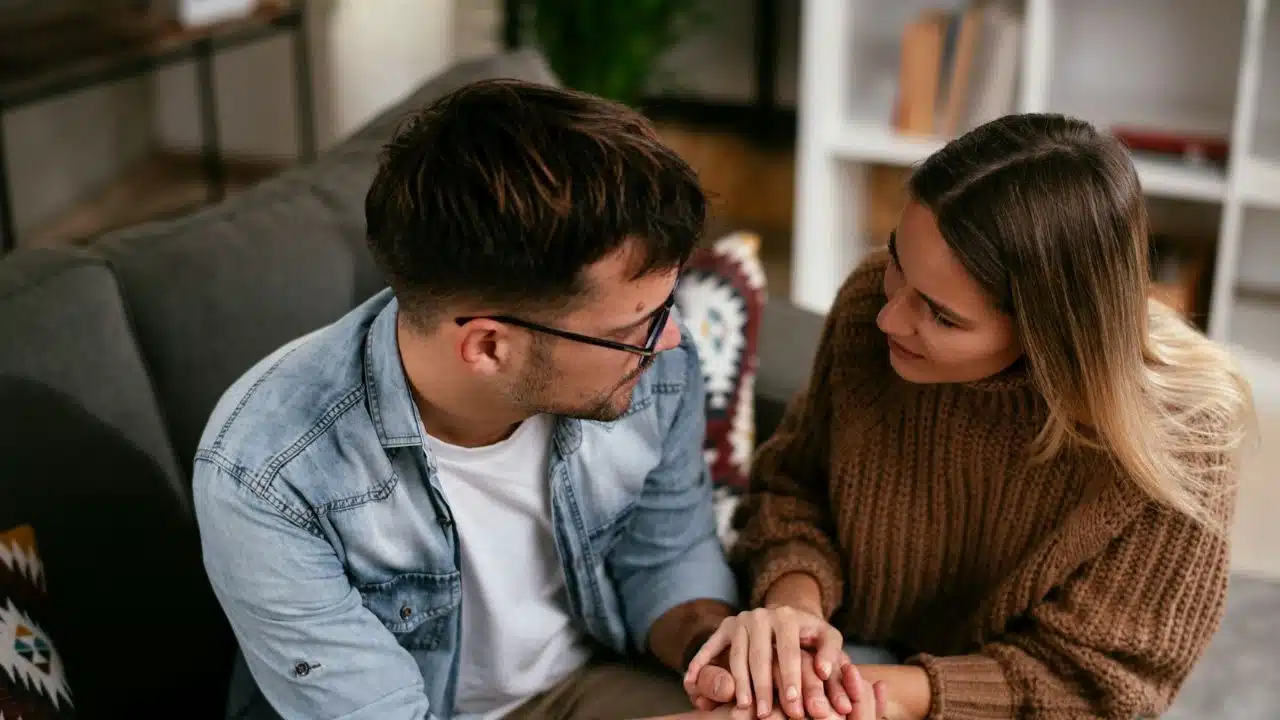 A man and woman sit closely on a couch, holding hands and looking at each other.