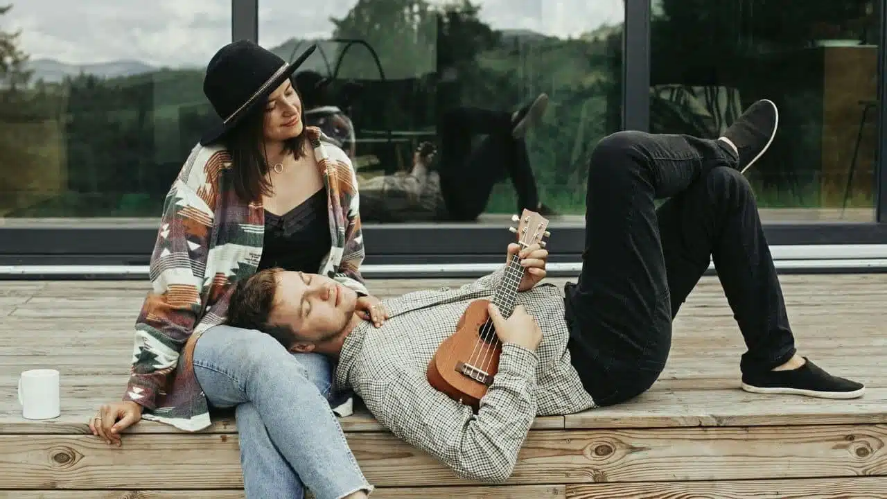 A man rests his head on a seated woman’s lap while holding a small ukulele.