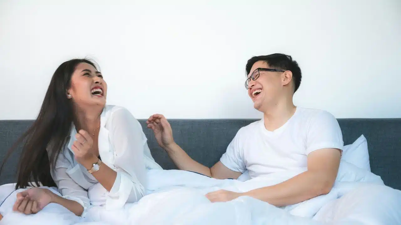 A man and woman sit on a bed laughing heartily while looking at each other.