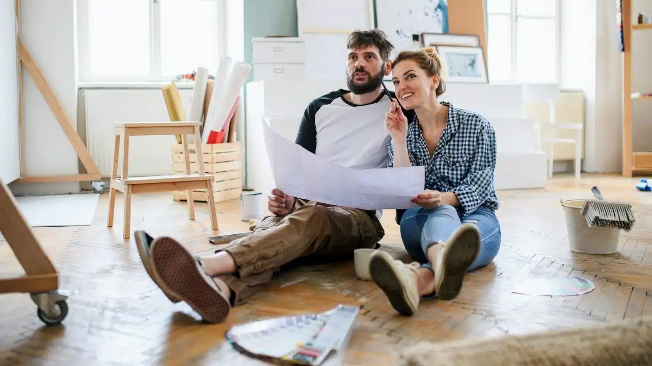 A man and woman sit on a wooden floor holding a large paper blueprint together.