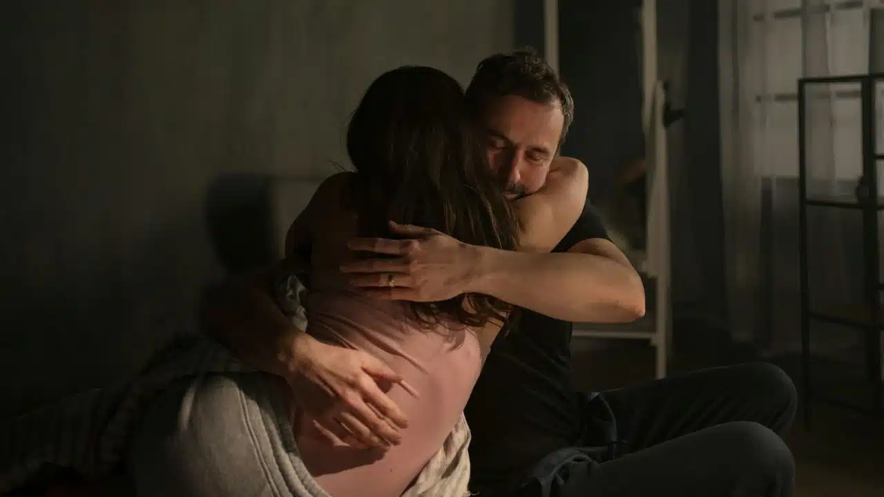 A man and woman sit on a floor in a dimly lit room, hugging each other.
