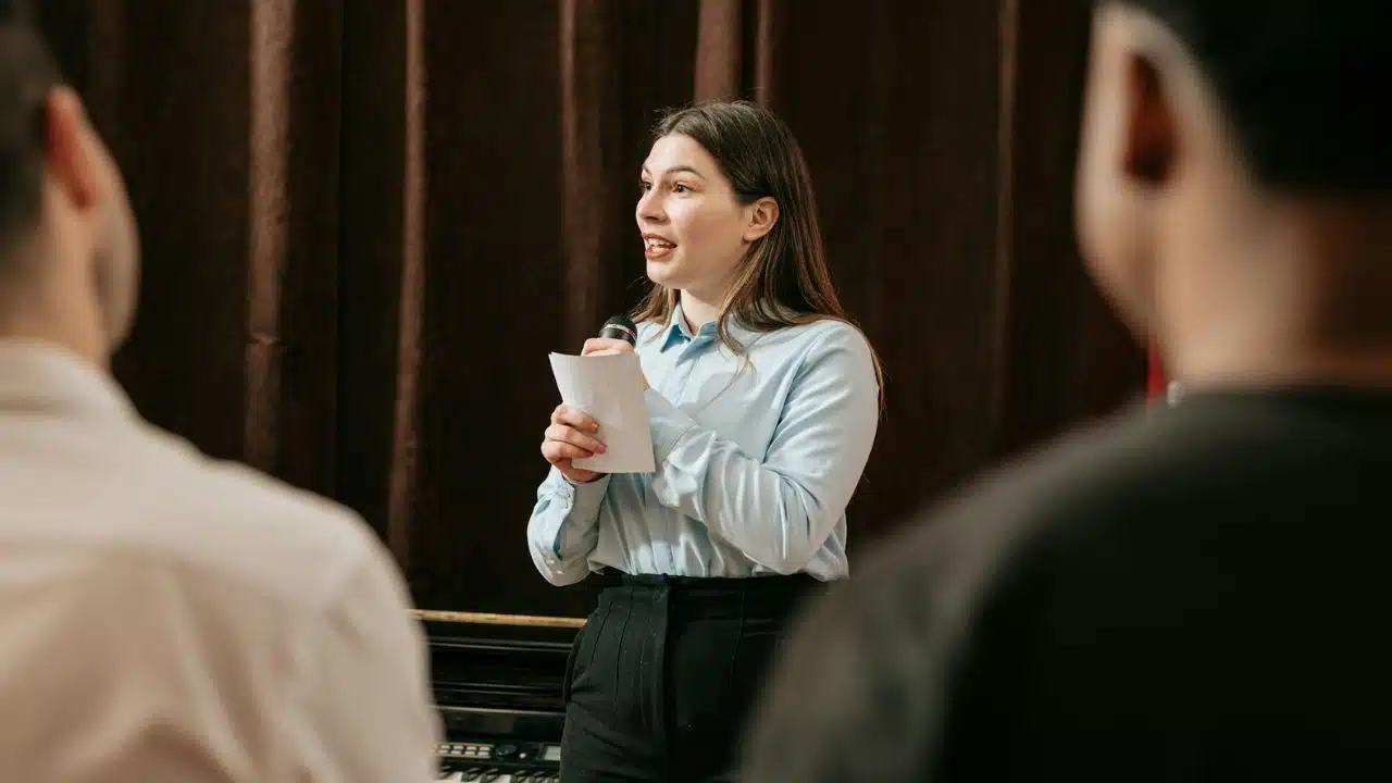 A woman stands holding a microphone and a piece of paper while speaking to others.