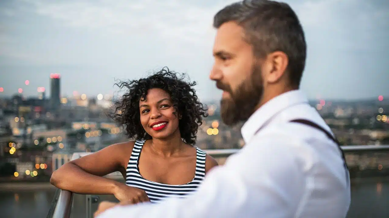 A woman in a striped top smiles at a man on a rooftop at dusk.