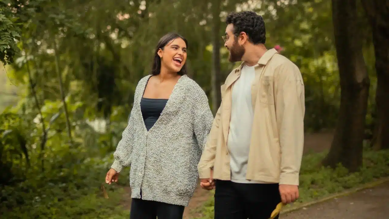 A man and woman walk through a wooded area holding hands while looking at each other.