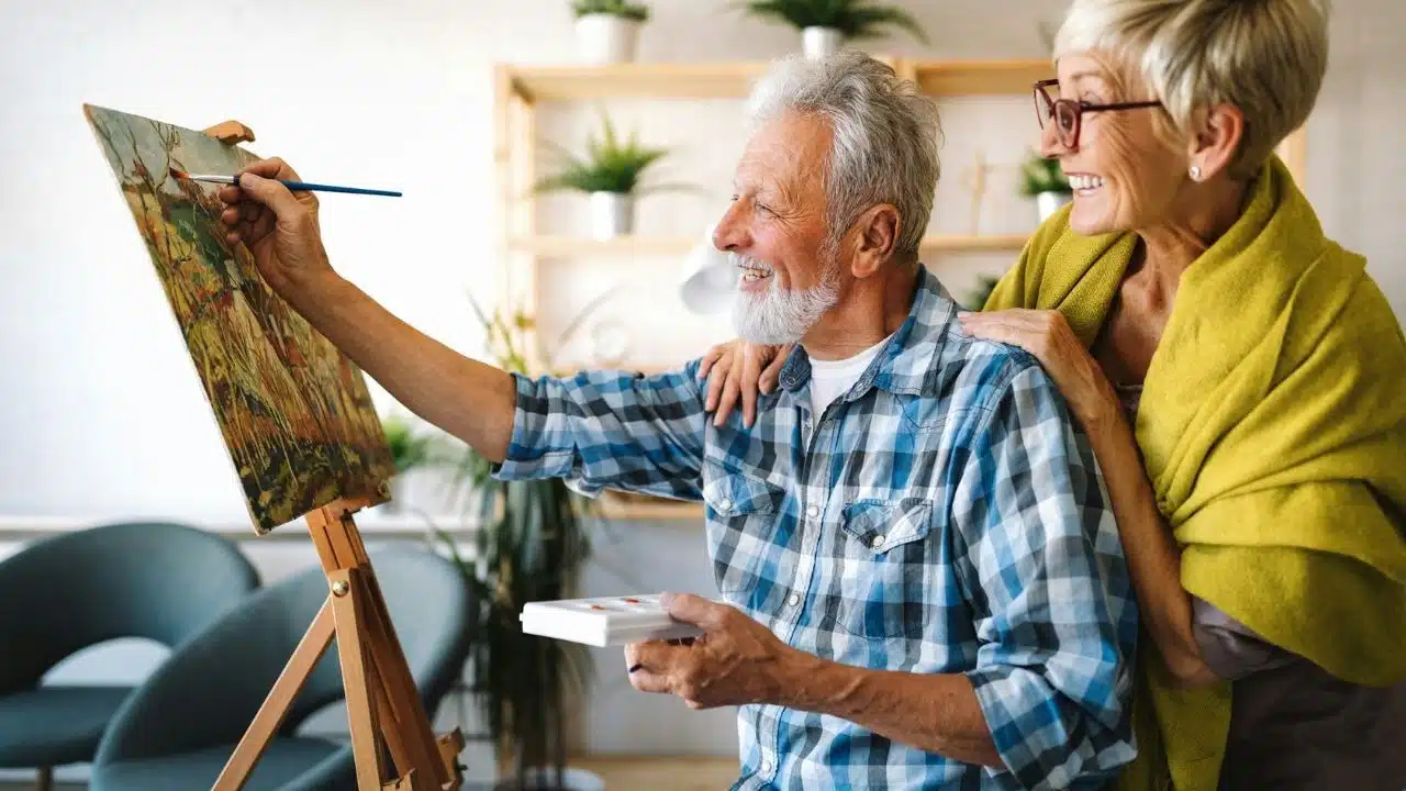 An older man paints on a canvas while an older woman smiles behind him.