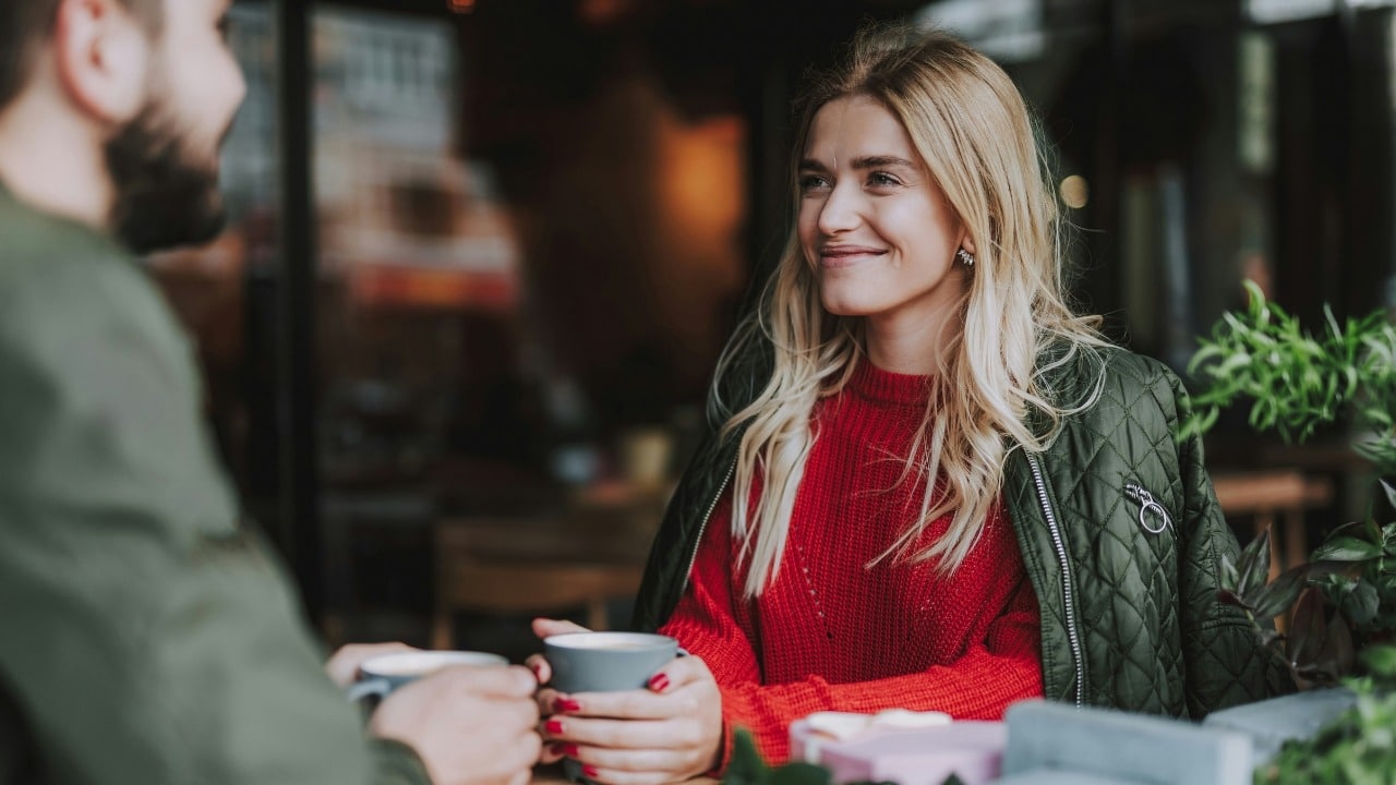 A blonde woman in a red sweater and green jacket smiles while holding a grey mug.